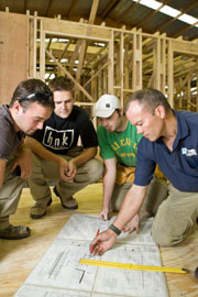 three studenst and a tutor on their haunches in front of wooden house frame they are looking at house plans 