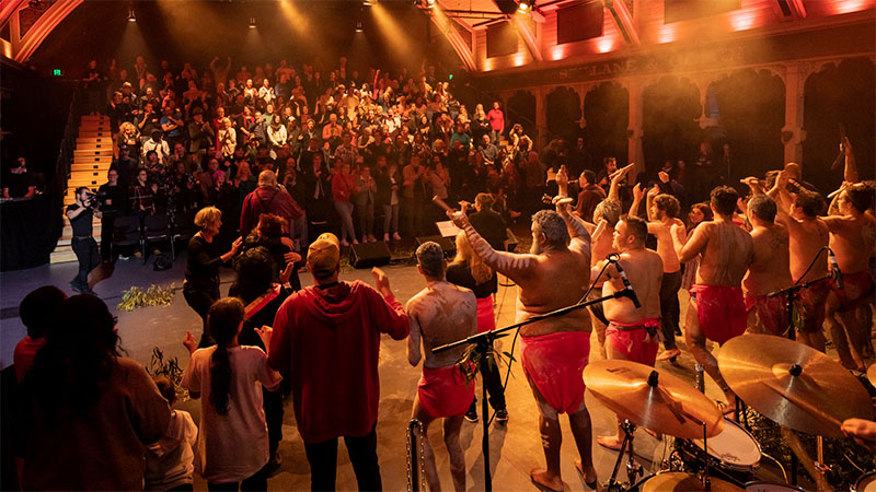 Aboriginal performers on a stage, from behind, with the audience in the background looking at the performance