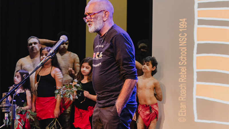 Professor Gary Foley on stage with a microphone, with Aboriginal performers in the background
