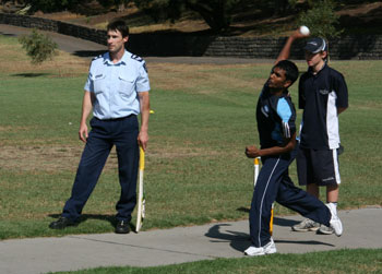 A student bowlers sends one down as a police batter stands by