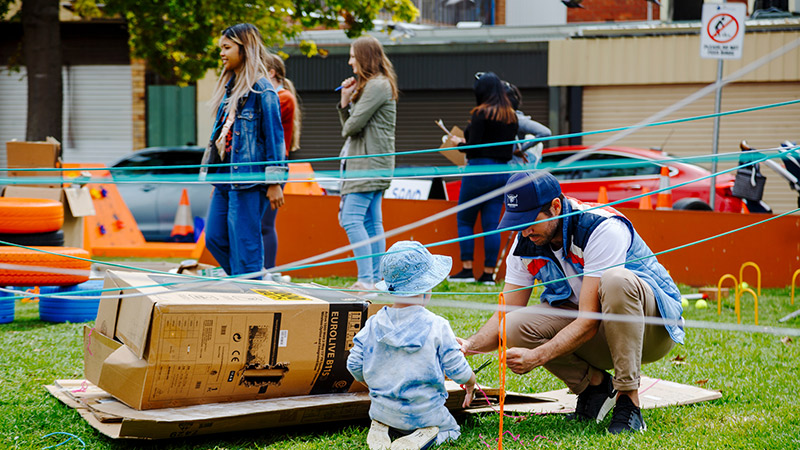 Child plays with their father outdoors in Footscray using string and boxes