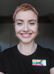 A young woman with short, pink hair, nose rings and a rainbow badge smiles at the camera