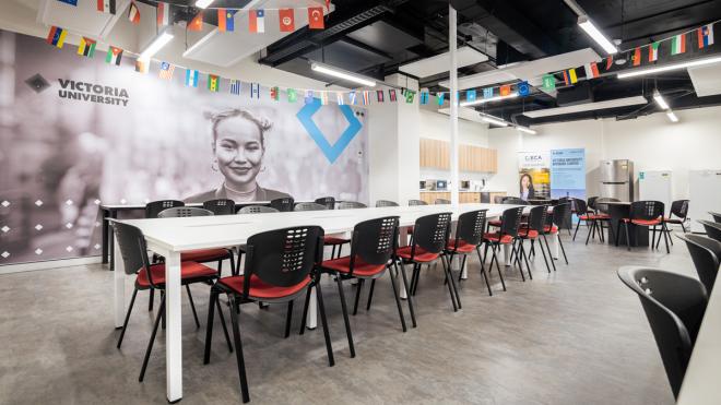Our break room on the ground floor includes a kitchenette for staff and students to grab a bite to eat between classes. Large room with chairs and a long table. In the background is a fridge and kitchen, with several Victoria University banners surrounding it. Several different countries' flags hang from the ceiling.