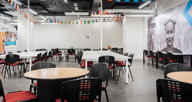  A large seating area with a combination of long, rectangular tables and small circle tables. Chairs surround the tables. Different countries' flags hang from the ceiling. Victoria University banner hangs in the background.