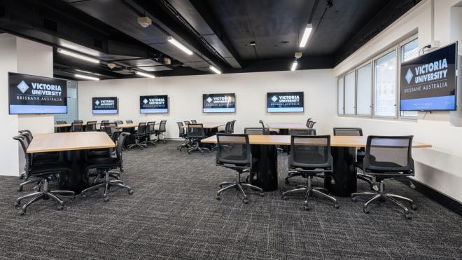 Our classrooms on campus offer space for collaborative learning and comfortable seating. A large room with long tables and computer chairs surrounding them. Each table has a computer screen next to it that is mounted to the wall.
