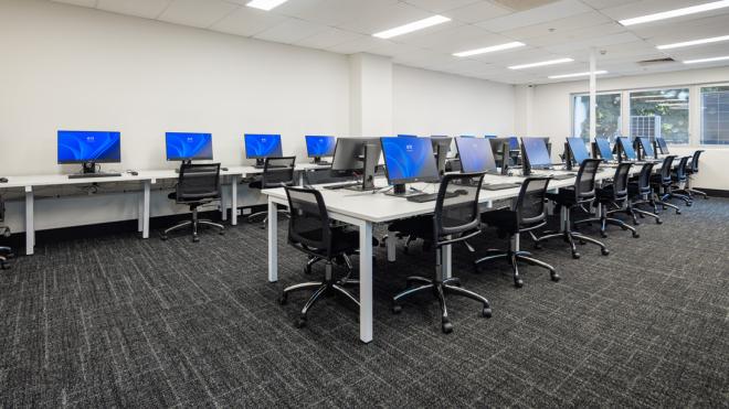 Our computer lab on Level 1 helps students access the technology they need for their studies. A room with computers. In the middle of the room is a long table with computers on it and chairs surrounding it. At the far wall is another table with computers and chairs.
