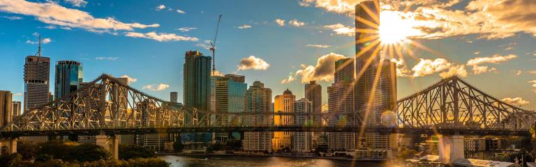  Brisbane skyline with bright sun peaking otu behind a tall building, with beautiful blue sky and a few clouds