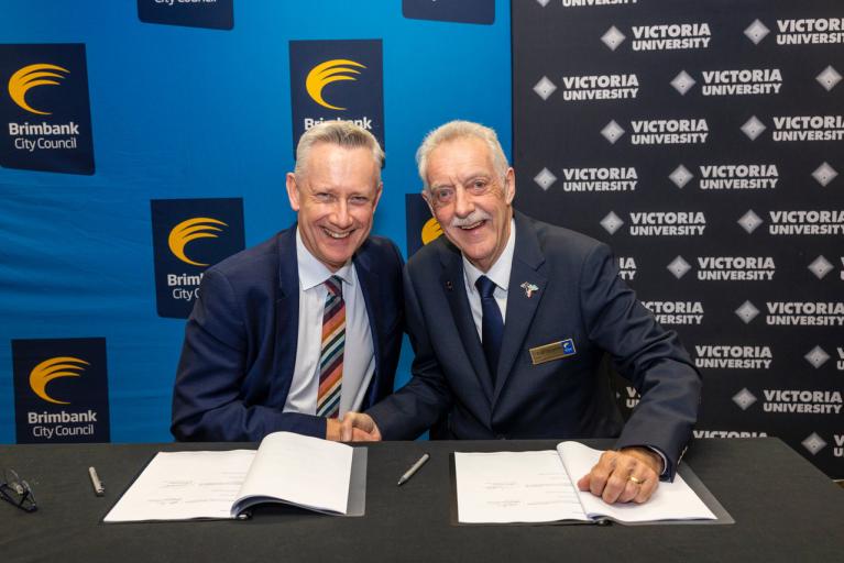 Two older white men in suits shake hands at a desk with signed forms in front of Brimbank Council and Victoria University logos
