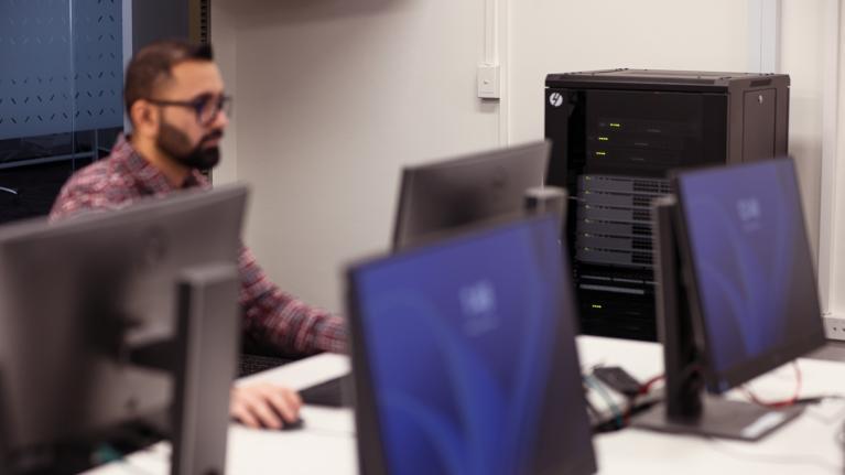  A student sitting at a computer. Three other computers are on the same desk. In the background is a large black computer.