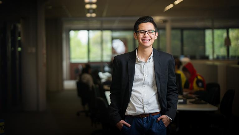  Sam posing casually, smiling with hand in pockets, in front of students at a computer hub