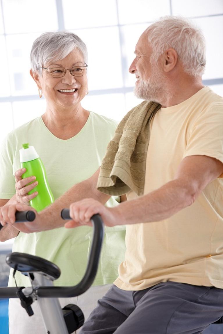  elderly man on an exercise bike with elderly woman smiling next to him holding a water bottle