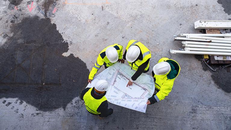  A top view of group of engineers with blueprints standing on construction site.