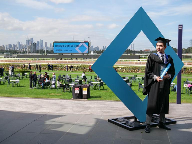  graduate posing in front of VU diamond symbol, at Flemington Racecourse graduation