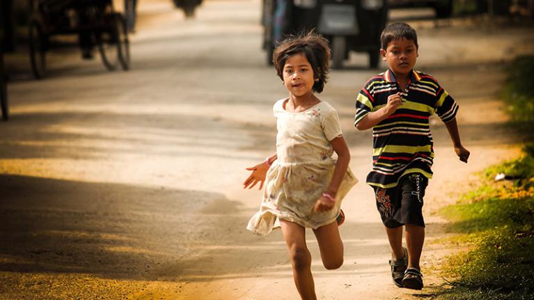 Two Indian children running on a dirt road, with vehicles in the background