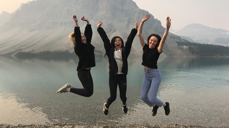 3 young women jump joyously in front of a beautiful lake and mountain