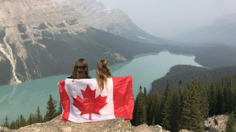 two female students look out over a vast lake and mountains, drapped in a Canadian flag