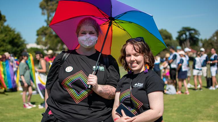  Two people wearing VU pride shirts on a sunny oval. One person is holding a rainbow umbrella. 