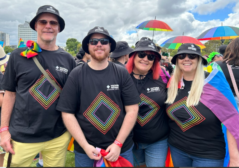 A group of smiling people in rainbow clothes with Victoria University logos