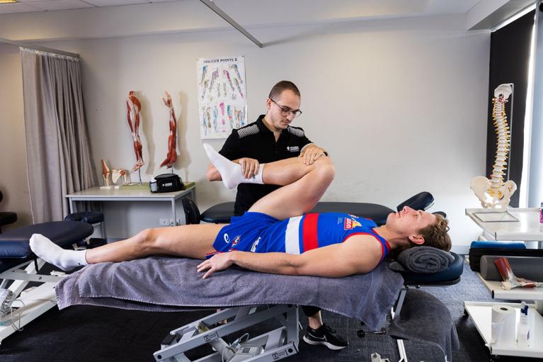 In a massage room with anatomical models, a young man manipulates the leg of another man in an red and blue sport uniform, on a massage table