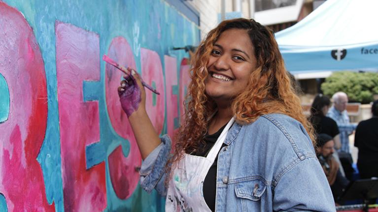  A smiling student is wearing an apron. They are standing, painbrush poised, at a green and pink mural saying 'RESPECT' in large letters. mural 