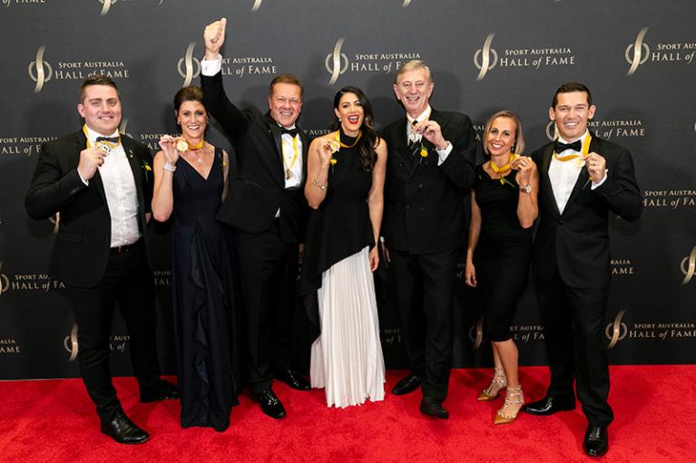 Seven men and women, in formal dress, looking happy and showing gold medals on a red carpet