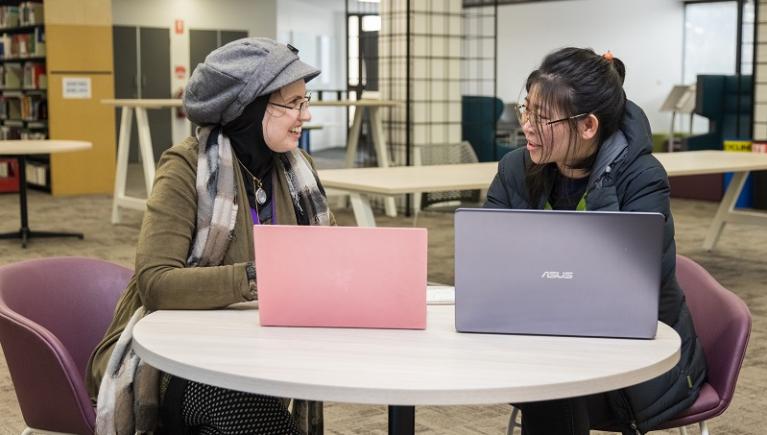 Two students, multicultural, chat over laptops in the library