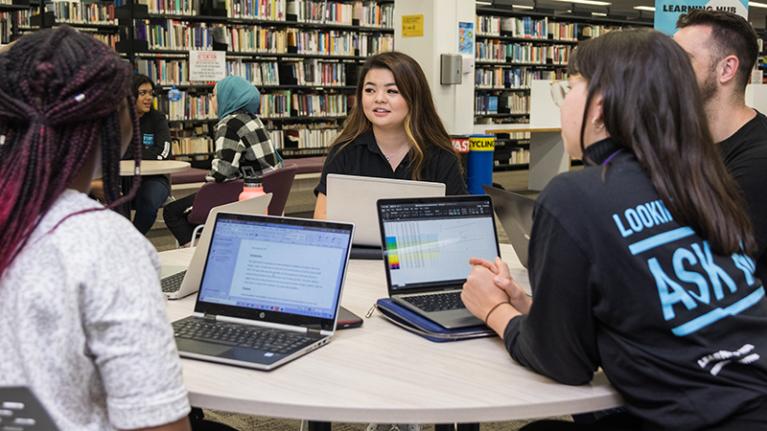 Group of students, multicultural, some wearing mentor shirts, converse around a library table with laptops