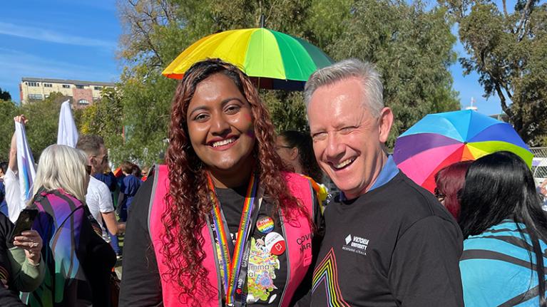  Vice-Chancellor Adam Shoemaker with student at Midsumma Pride March. Both have big smiles. Rainbow umbrellas are in the background. 