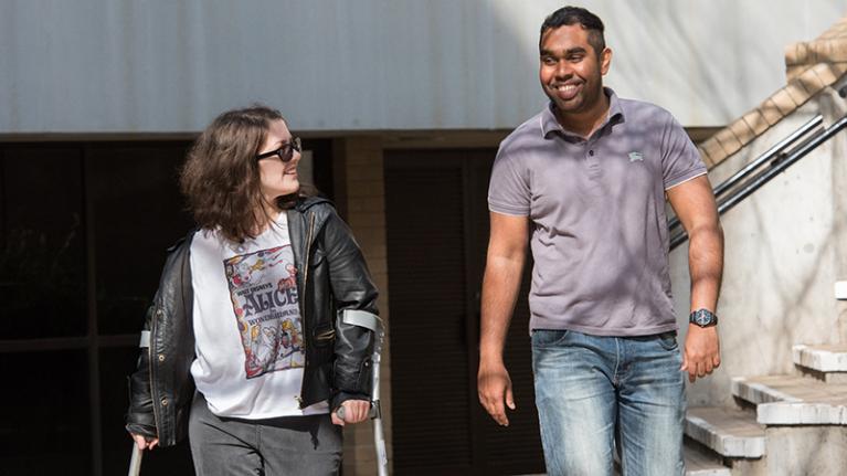  Two people walking and smiling. One of them is using crutches. 
