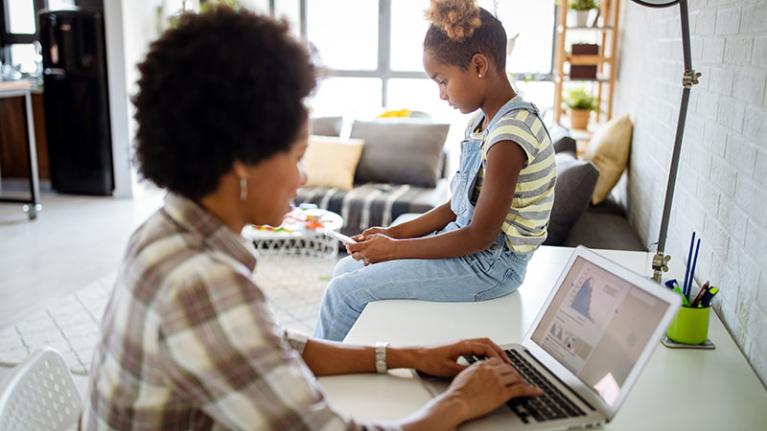  A woman works on a laptop as a child sits next to her on a desk.