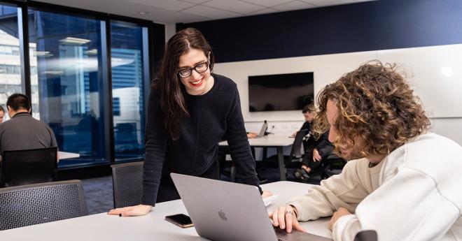 Young man at a desk in a modern classroom shows a smiling woman in glasses something on a laptop