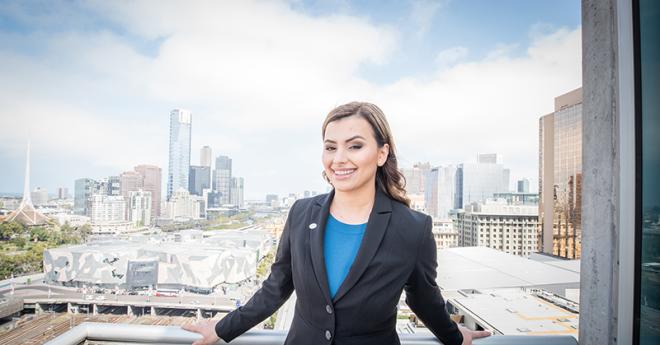 A young woman in professional dress stands on a city balcony with a cityscape behind her
