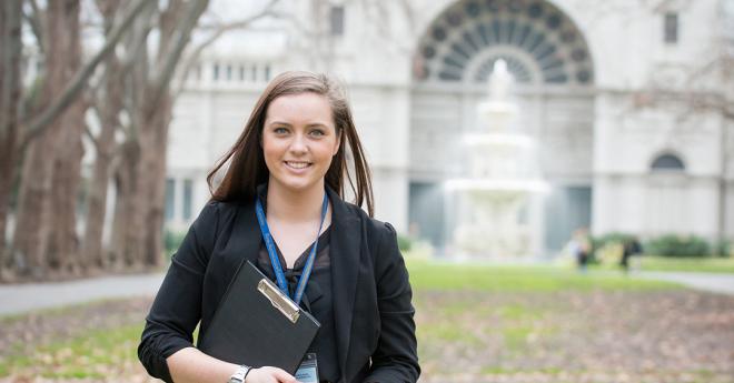 A young woman with a clipboard strids across and autumn lawn in front of an elegant building