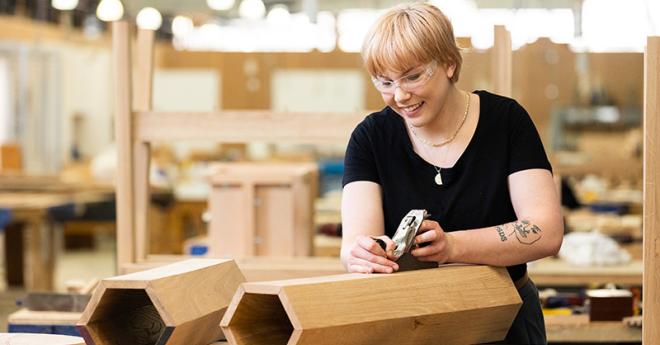  A young woman smiles as she planes a hexagonal woodwork creation in a large workshop