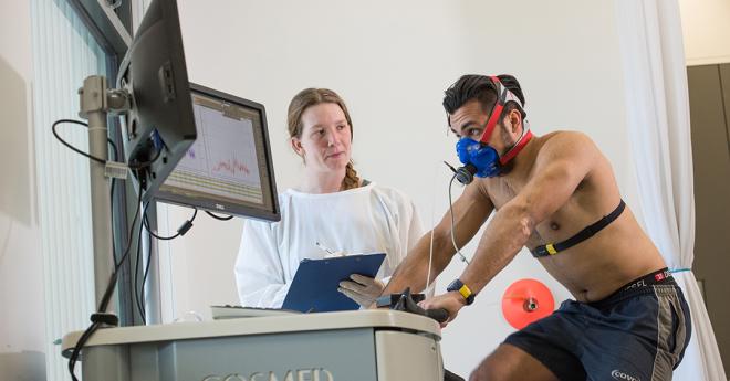 man hooked up to monitoring equipment rides an exericise bike, watched by a woman in a lab coat