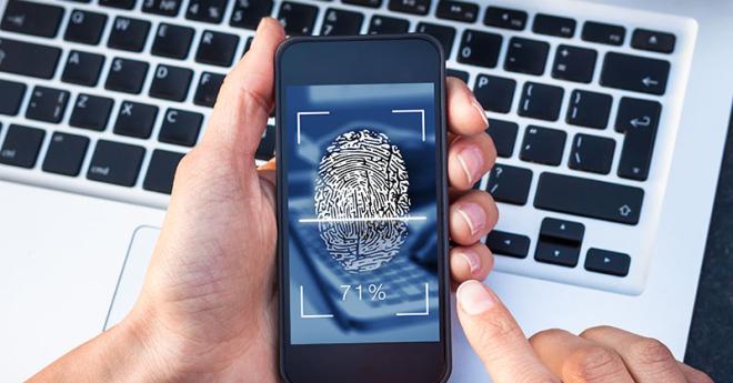  Hand holding a mobile phone in front of a laptop, with a security fingerprint icon on it.