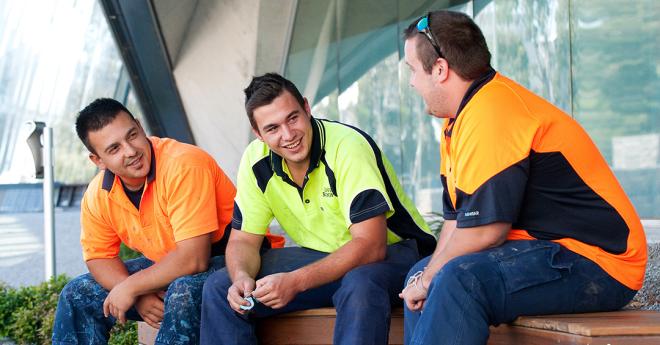 three young men in trades uniforms chat outside a modern campus building