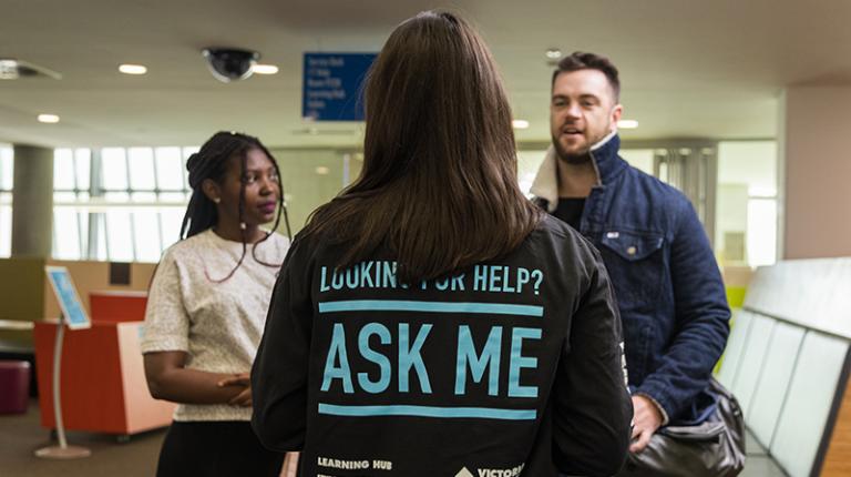 Back of student showing 'Ask me' written on shirt; talking to white male and African-Australian students
