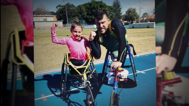 young girl using a 3-wheel racing wheelchair, hand raised in victory, with an adult wheelchair athlete