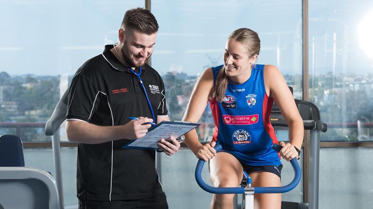A woman in Western Bulldogs attire on an exercise bike, with a VU staff member standing beside her, with a clipboard and stethoscope