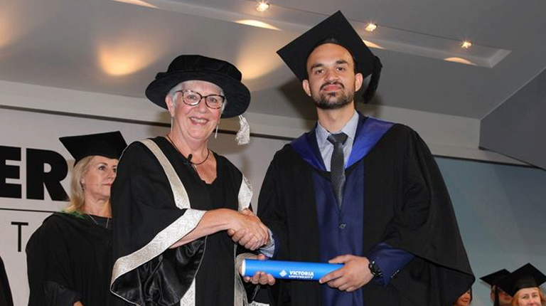  undergraduate receiving his testamur and shaking hands on stage at graduation ceremony