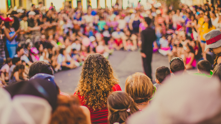 Crowds standing and sitting in a circle around a figure, blurred in the background.