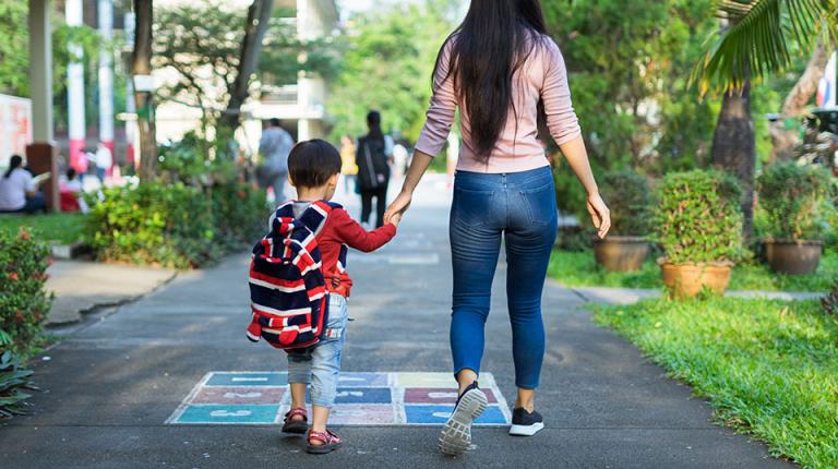 A young boy wearing a backpack walking to school holding his mother's hand.