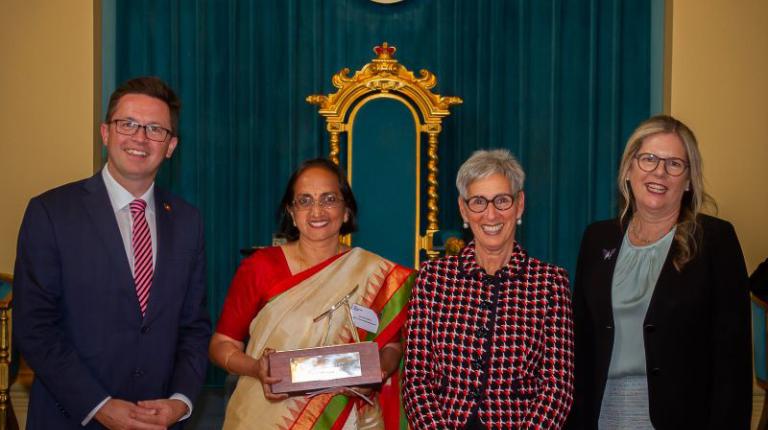 Victorian MP Anthony Carbines; Jaya Manchikanti; Governor of Victoria, Linda Dessau; and Penny Fowler, granddaughter of Elisabeth Murdoch Group of people stand in front of colourful curtain, including woman in traditional Indian dress, holding award