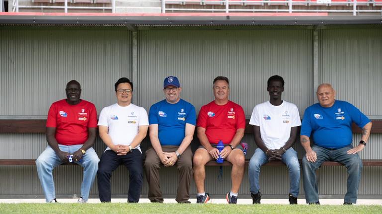 Group of 6 men from diverse ethnic backgrounds smile at the camera from a footy bench, wearing red white and blue