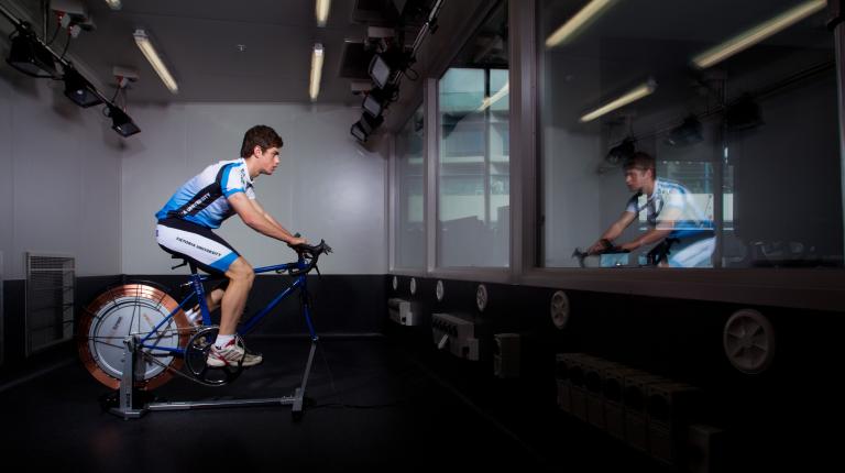 VU’s College of Sport and Exercise Science and the Institute for Health and Sport are ranked 9th in the world. A male-presenting person riding an exercise bike indoors in a gym.