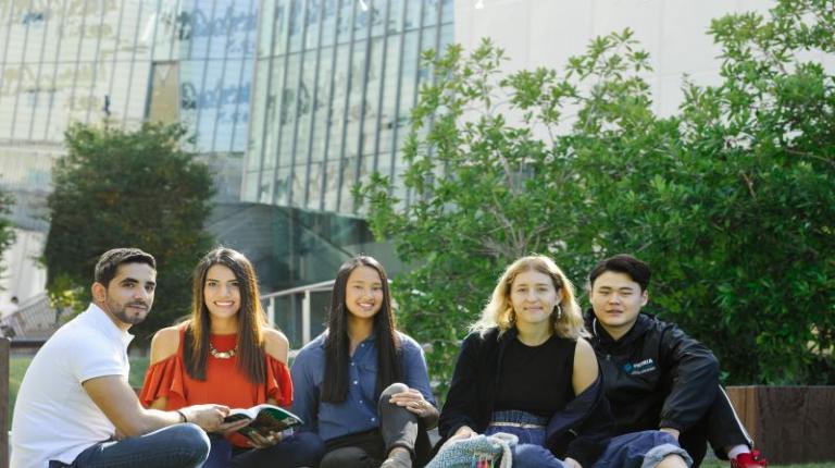 Five diverse, smiling students sitting on the grass at Footscray Park Campus