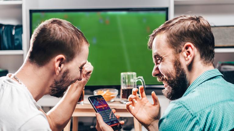 Two friends excitedly checking their sports betting app on their phone, as they watch a soccer match together