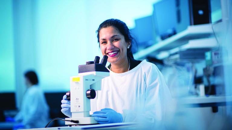   	A female researcher in lab-coat and gloves at a microscope in a lab. 