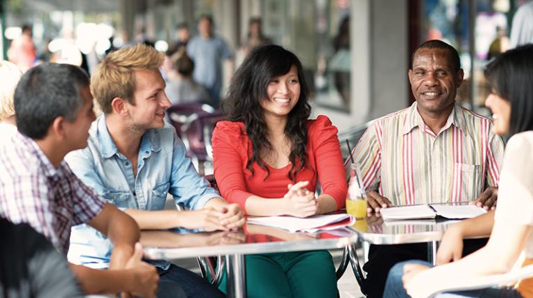 Five international students sitting outside in the street at a table talking. 